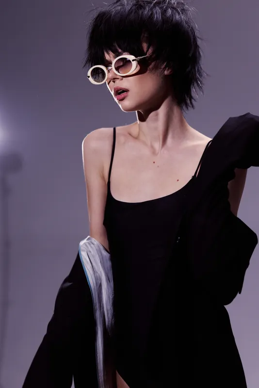Stylish woman with short hair wearing black outfit and round sunglasses posing indoors with dramatic lighting and shadow background