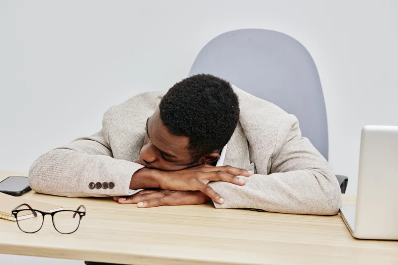 Relaxed black man asleep at office desk wearing beige blazer with closed eyes in a modern workspace with laptop and glasses isolated on plain background. People concept