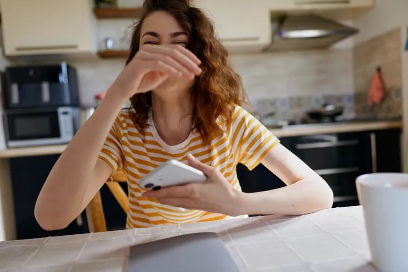 Young woman with brown hair sits at a kitchen table, laughing into her hand while scrolling on a smartphone in a casual striped tshirt, bright morning light creating a cozy home atmosphere.