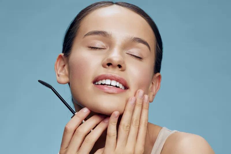 Young woman applying skincare with a brush, showcasing healthy skin and radiant smile against a soft blue background, reflecting self care and beauty rituals
