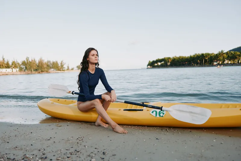 Person sits beside a yellow kayak on a sandy beach, calm water nearby, reflective mood, casual long sleeve top, soft light, serene outdoor scene inviting gentle exploration.