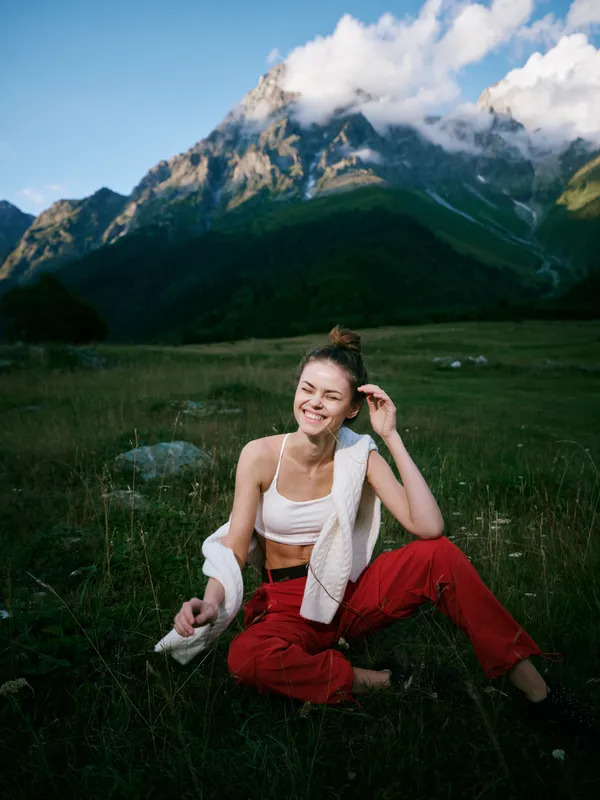 Outdoor mountain fitness landscape with a smiling athlete resting on a meadow, towel over shoulder, casual sportwear, against rugged peaks and a bright sky after activity