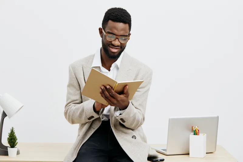 Confident young man with dark skin, glasses, and curly hair dressed in smart casual, reading a notebook while sitting at a modern office desk with a laptop, stationery, and a potted plant, in bright m