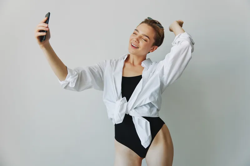 Подросток-блогерша фотографирует и снимает себя на видео на телефон для контента в социальных сетях, делает селфи с улыбкой на фоне белой стены дома, фрилансер