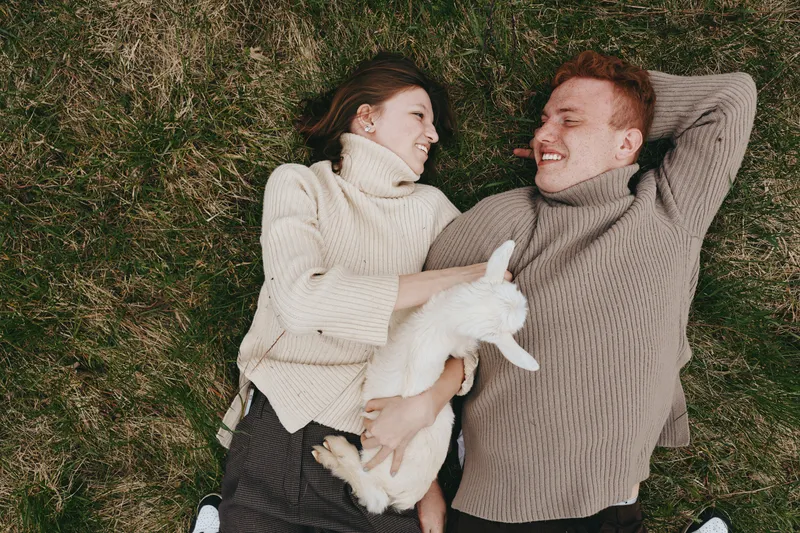 Cheerful couple outdoors, lying on grass, sharing a laugh in cozy sweaters, sunlit moment, relaxed pose, gentle breeze, natural tones, spontaneous affection, simple happiness in a serene park setting