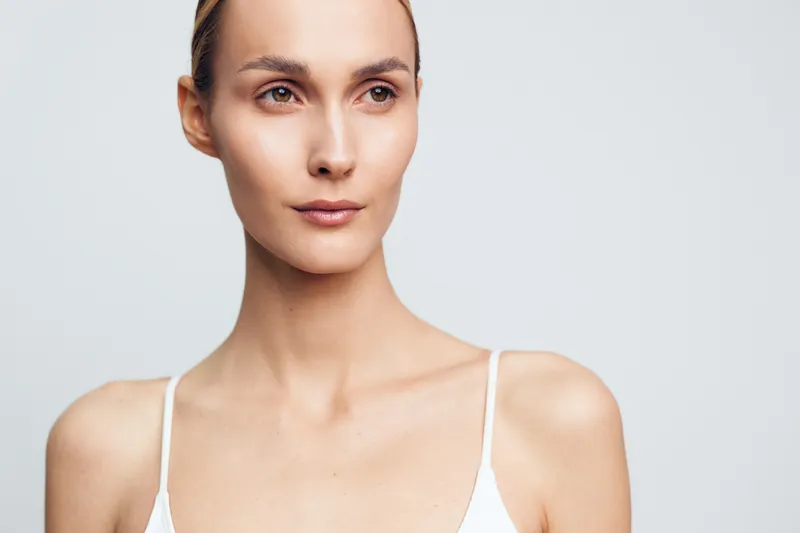 Portrait of a calm young woman with natural skin wearing a white tank top looking aside on a light gray background with soft lighting and minimal makeup.