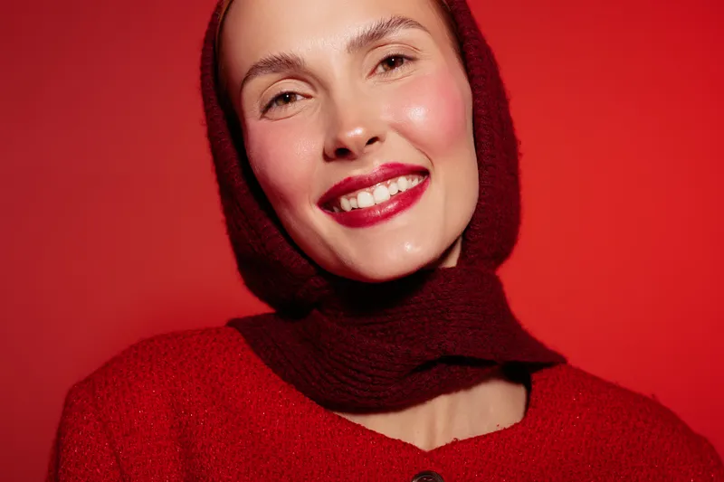 Smiling woman with red lipstick and knitted scarf poses against red background. Close-up portrait with warm lighting highlights natural skin texture and joyful expression. Fashion concept.