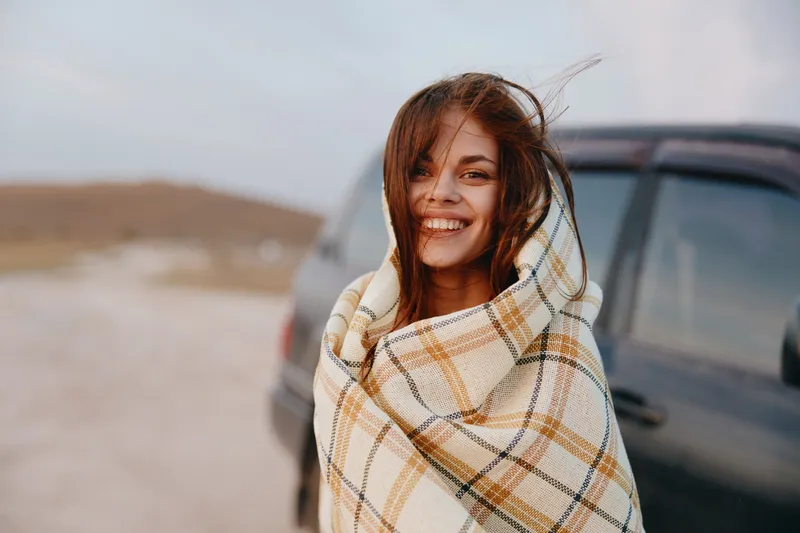 Улыбающаяся красавица, укутанная в плед, стоит на обочине дороги перед машиной в путешествии, полном приключений