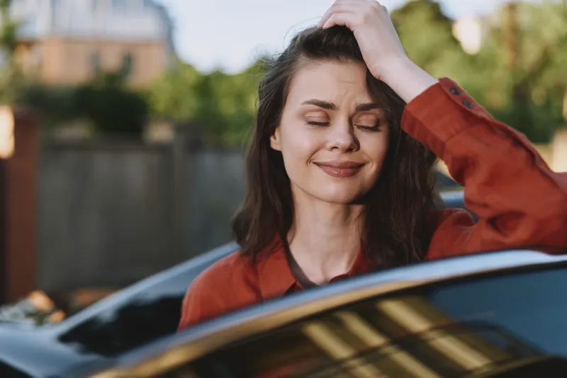 Young woman with dark hair smiles softly as she brushes her hand through her hair while leaning against a sleek car on a bright, sunlit day