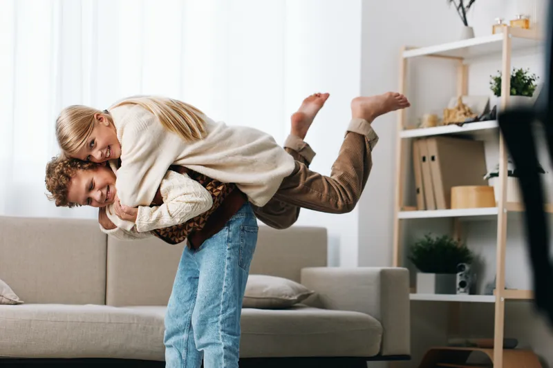A joyful couple playfully wrestles on a cozy living room couch, laughter lighting their faces as a carefree moment freezes in bright natural light.