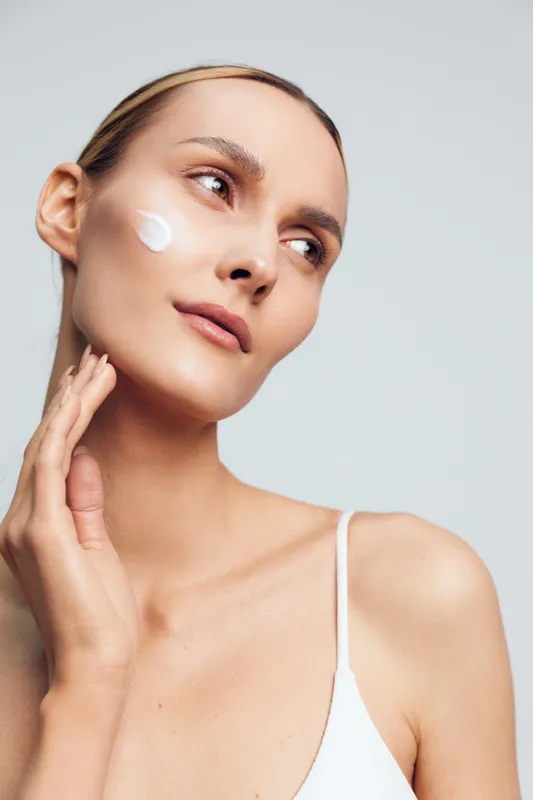 Skincare beauty portrait of a woman in studio lighting, gently touching her face, applying cream to the cheek, calm wellness expression, smooth skin, minimalist background