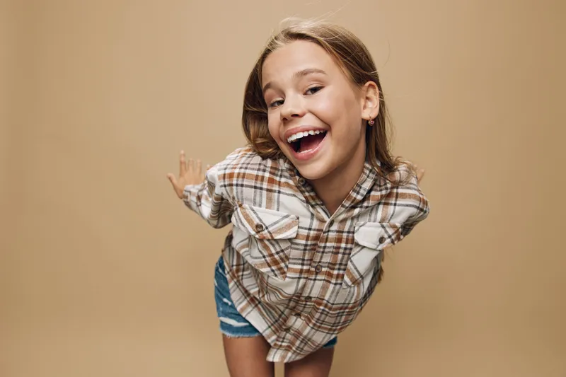 Girl smiling and posing with arms behind back in plaid shirt and denim shorts standing on beige background. Happy child with casual outfit and playful expression in studio.