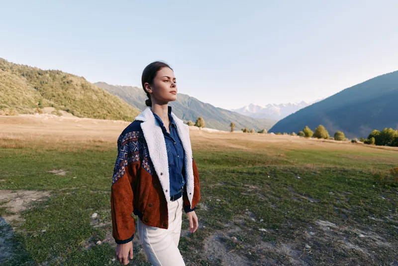 Woman nature mountains outdoors meadow walking portrait of a young woman in cozy jacket strolling across grassy valley at golden hour, calm gaze and scenic landscape behind her.