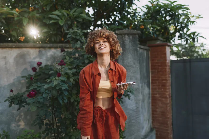 A woman outdoors in a sunlit garden wears an orange outfit. A candid lifestyle portrait capturing confidence, warmth, and carefree summer vibes with natural light