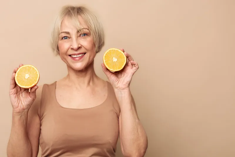 Smiling mature woman holds two halves of fresh orange near her face, wearing beige tank top. Healthy lifestyle, vitamin C, citrus fruit, wellness concept on neutral background.