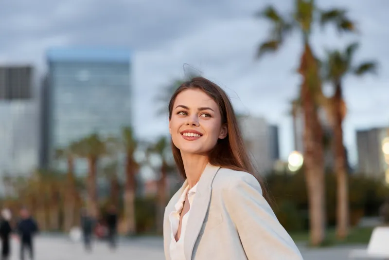 confident young woman in light blazer smiling outdoors with urban blurred background and palm trees, business style, natural light, fresh atmosphere