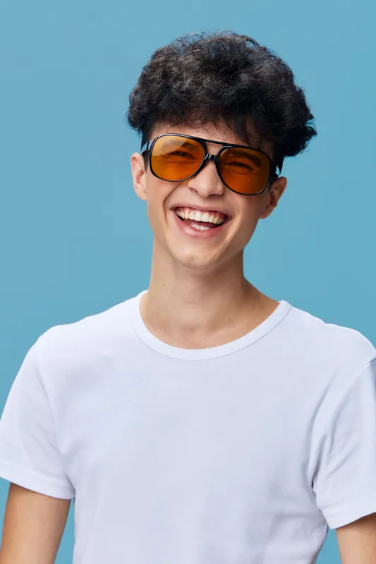 Smiling man wearing orange sunglasses and a white t-shirt, expressing happiness with curly hair against a plain blue background in bright studio lighting.