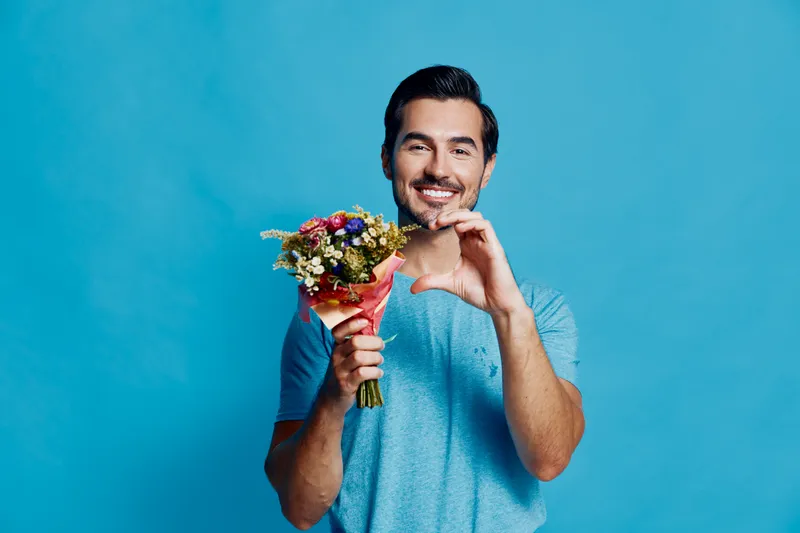 Full body smiling happy cheerful young man holding a bouquet of flowers, making a heart shape with his hands, set against a bright blue background.