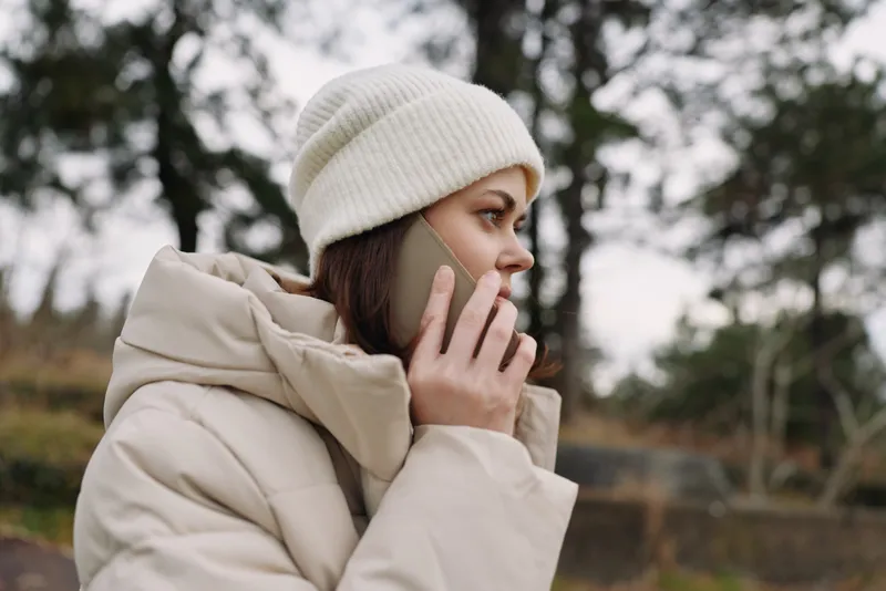 Woman in a quilted beige coat and white beanie talks on a mobile phone outdoors, winter scenery with bare trees under gray skies, creating a calm candid moment. The scene emphasizes street fashion,