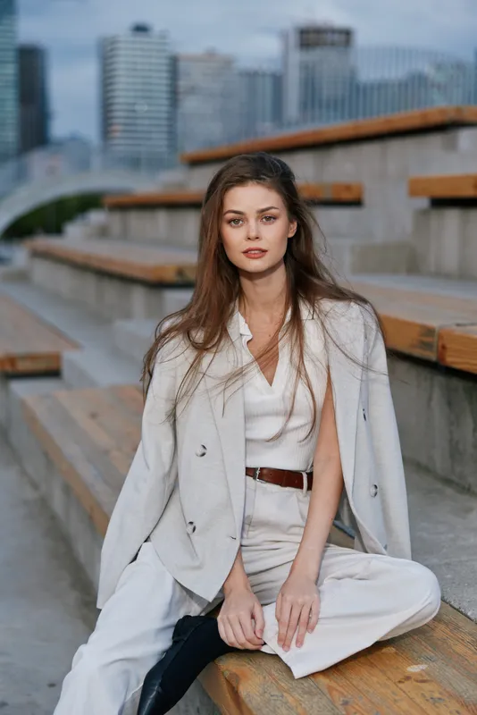 Young woman in beige business casual outfit sitting on wooden steps with urban cityscape background, natural light and confident expression