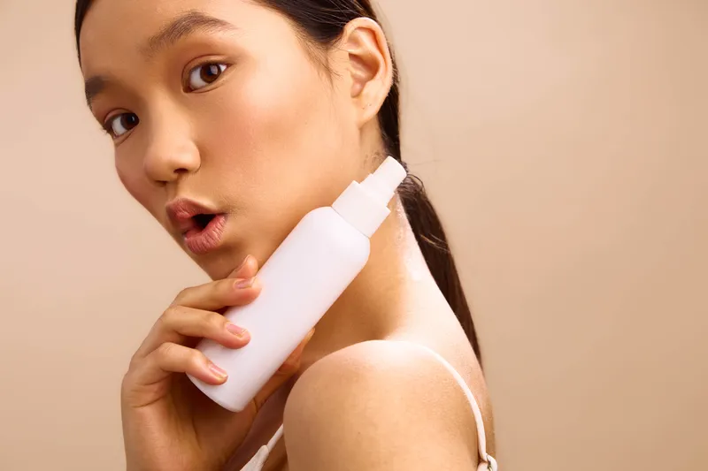 asian woman holds a spray bottle near her shoulder in a soft studio, presenting a beauty product with a calm gaze and gentle glow, creating a clean, modern and inviting aesthetic.