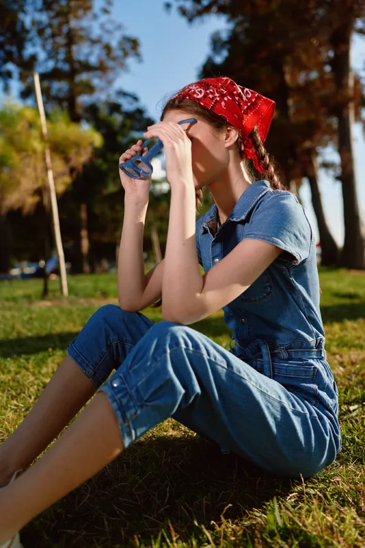Lifestyle woman in modern boho-western denim street style sitting outdoors drinking from a cup with film color tones and natural sunlight in a park setting.