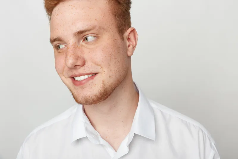 Young redhead man with freckles smiles looking sideways wearing white shirt on light background. Concept of casual style, happiness, and natural male portrait in studio lighting.