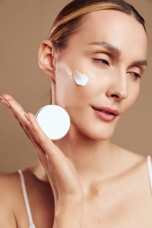 Young woman with smooth skin applies white cream on cheek holding cosmetic jar close to face demonstrating skincare routine and healthy beauty concept on beige background.