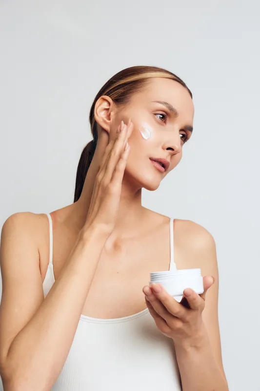 A young woman in a white tank top applies moisturizer cream to her face with her fingers against a plain background. Concept for skincare, beauty routine, and healthy skin care.