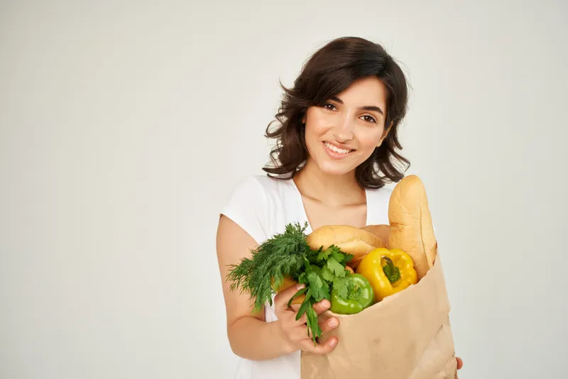 Весёлая женщина с пакетом продуктов в супермаркете на светлом фоне