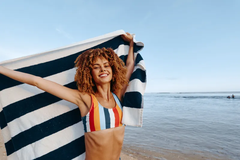 A young adult with curly hair stands on a sunny beach, holding a striped towel above shoulders, wearing a colorful bikini, radiating a carefree summer moment.