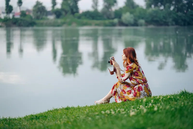 Женщина на открытом воздухе в платье с фотоаппаратом в руках на берегу озера, отдых