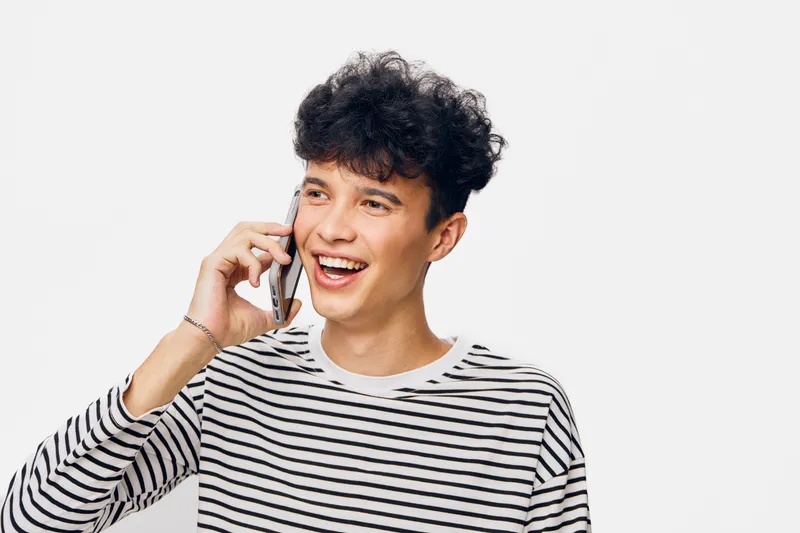 smiling man talking on phone wearing striped sweater with curly hair and bright expression against white background in casual indoor setting