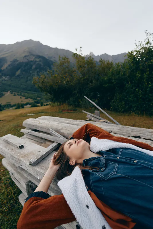 Woman outdoors on wooden pallets in a meadow near mountains, nature portrait in countryside, a moment of relaxation. Young woman in denim and shearling jacket lying peacefully.