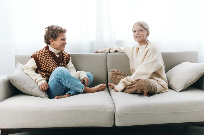 Two children sit on a light sofa, sharing a friendly conversation in a cozy living room. They wear knitted sweaters, relaxed posture, and soft natural light.