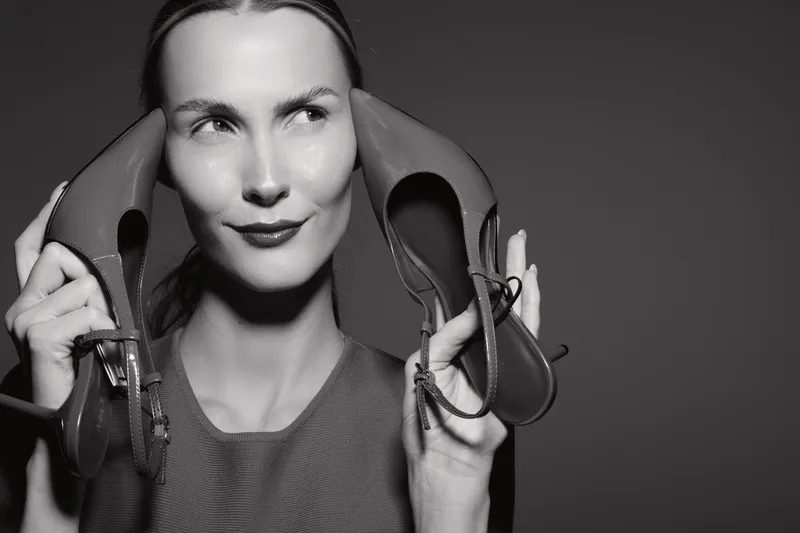 Portrait of a fair skinned Caucasian woman holding two high heel shoes near her face with a playful expression. Studio lighting highlights facial features and shoe details.