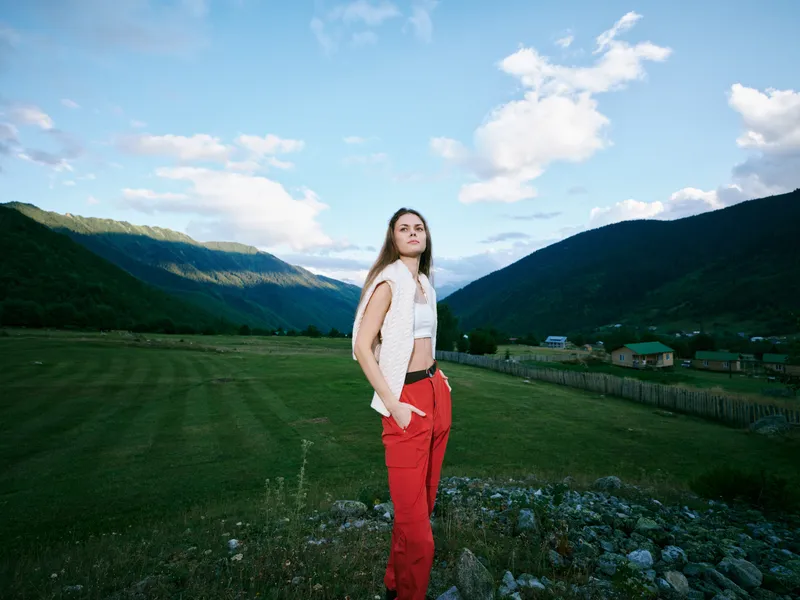 Woman standing in a grassy valley with mountains, clear blue sky, white top and red pants, confident pose, open space, natural light, fashion and travel vibe