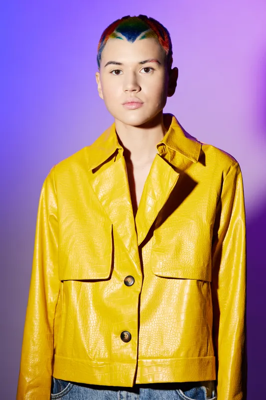 young woman with colorful short hair wearing bright yellow jacket stands confidently against vibrant purple background in contemporary fashion portrait