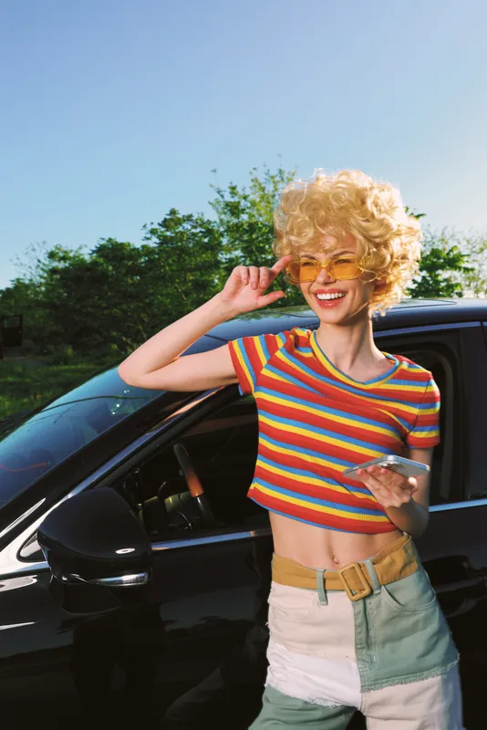 lifestyle, woman, fashion, retro, colorful, sunshine, outdoor, casual, smile, sunglasses. Smiling woman with blonde curls wearing a striped crop top and denim shorts beside a car outdoors, embodying