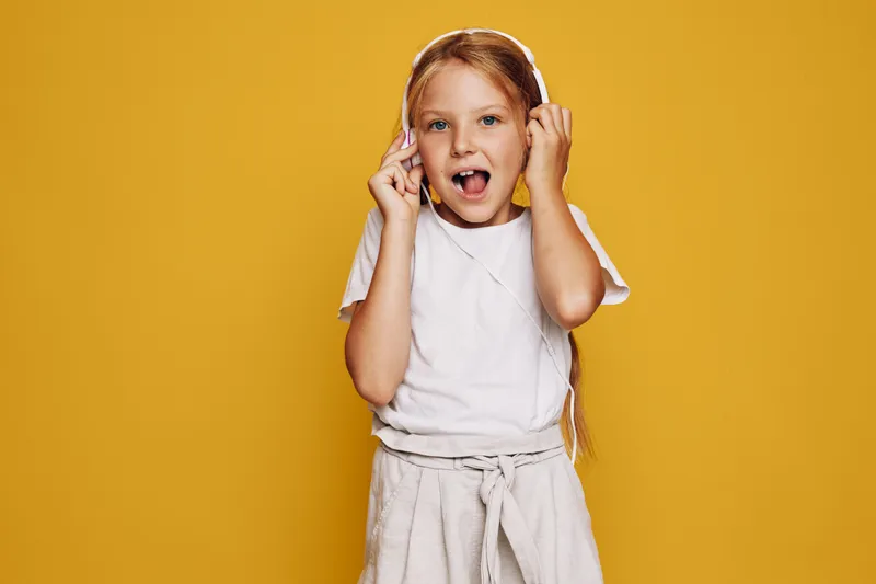 Young girl with long blonde hair wearing white headphones and casual clothes sings with open mouth against yellow background. Studio shot with bright lighting. Concept of music and childhood.