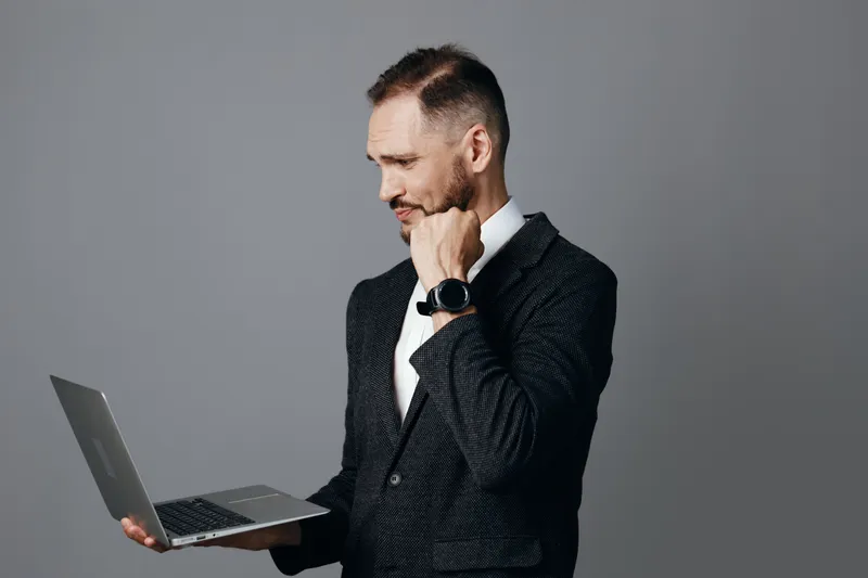 Professional man in a business suit stands against a colored isolated background, holding a laptop, conveying focus, confidence, and determination in a calm pose.