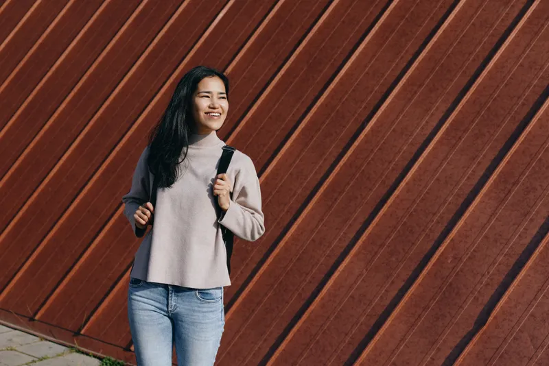 Smiling woman with backpack stands outdoors against a diagonal brown wall, showcasing casual street style, urban vibe, denim jeans and soft sweater in a candid lifestyle shot