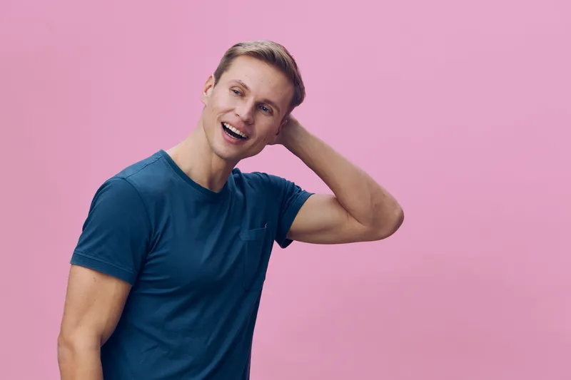 Smiling young man with short blonde hair wearing blue t-shirt on pink background, casual pose, cheerful expression, looking directly at camera, lifestyle shot, vibrant colors, studio portrait.