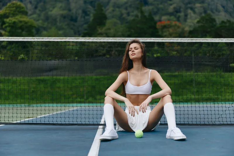 Young athletic woman in sports attire sitting on a tennis court, exuding confidence and relaxation amidst a lush green background and calm atmosphere