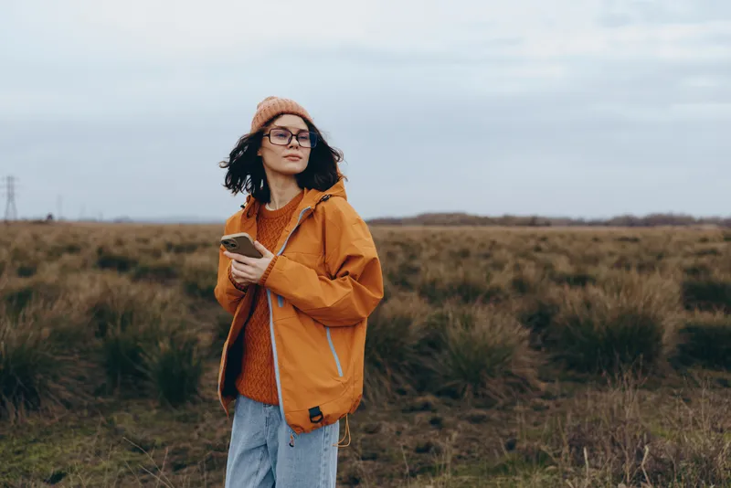 Smiling woman lifestyle outdoors in an autumn field, wearing orange jacket and beanie, with glasses, holding a smartphone, relaxed pose, overcast sky, natural light, casual travel