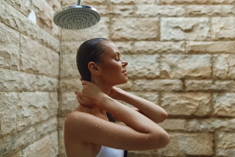 Woman taking outdoor shower with stone wall background, water droplets falling, serene expression, natural light highlighting her face and shoulders.