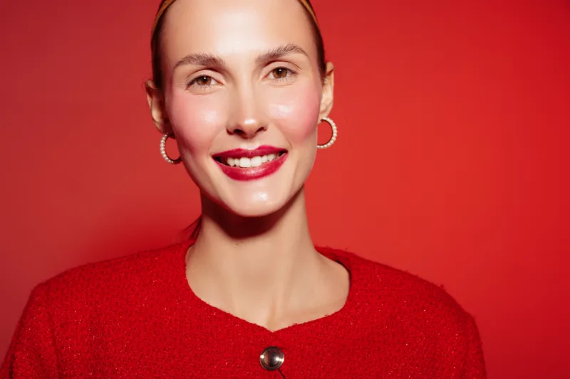 Smiling woman with red lipstick and hoop earrings wears red outfit against red background. Close-up portrait with natural skin glow and confident expression. Concept of beauty and fashion.