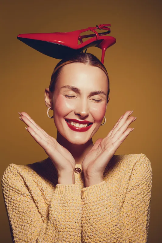 Smiling woman with closed eyes wears red high heel shoe on head, posing with hands near face. Fashion concept with playful mood and stylish accessories on warm background.