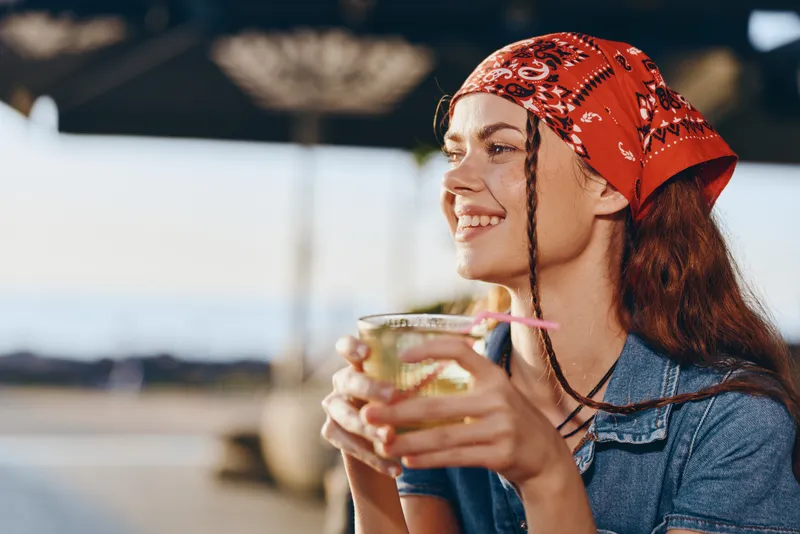Lifestyle woman in modern boho-western denim street style enjoying a drink, smiling brightly with film color tones under natural sunlight outdoors.