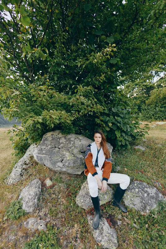 Woman sitting on rocks under a large tree in nature, wearing boots and casual jacket, outdoor portrait with relaxed pose, long hair, mossy stones and grassy meadow scenery.